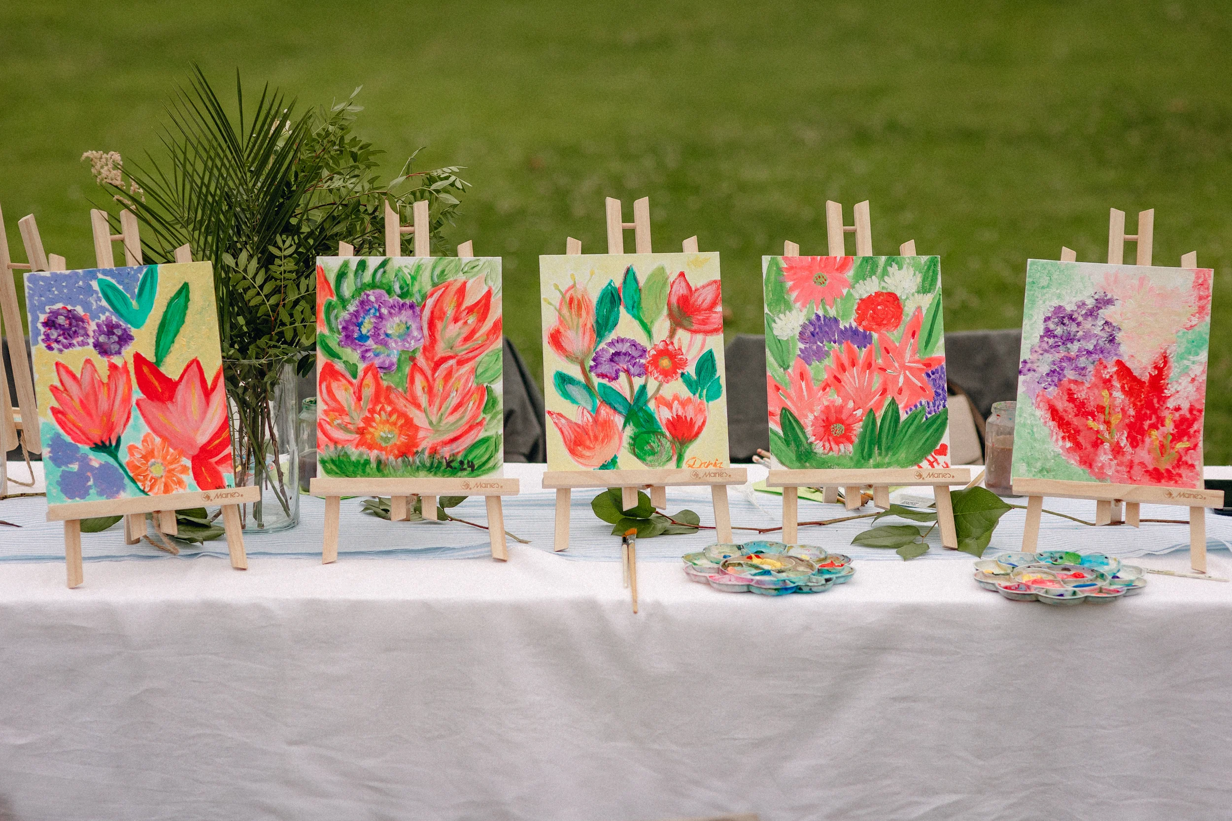 Atelier de peinture en plein air : plusieurs toiles florales exposées sur chevalets, posées sur une table blanche avec une touche végétale décorative